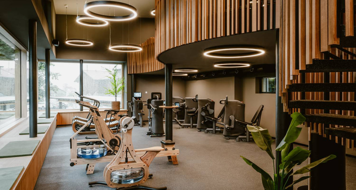 Hotel-Fitnessraum mit Rudergeräten aus Holz, Indoorbikes und runden Deckenleuchten vor einer Fensterfront